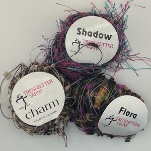 May include: Three skeins of Trendsetter Yarns in various colors. The skeins are labeled "Shadow", "Charm", and "Flora". The "Shadow" yarn is a mix of purple, pink, and teal. "Charm" is a mix of neutral tones. "Flora" is a mix of dark colors.