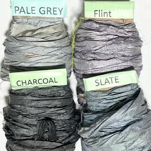 K&ouml;nnte beinhalten: Vier Spulen mit zerknittertem Seidenband in Graut&ouml;nen. Die obere linke Spule ist mit PALE GREY beschriftet, die obere rechte mit Flint, die untere linke mit CHARCOAL und die untere rechte mit SLATE. Jedes Band hat eine strukturierte Oberfl&auml;che.