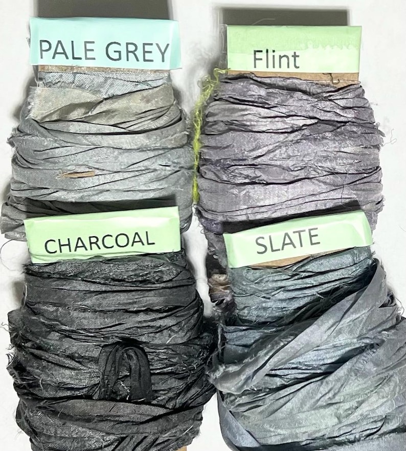 K&ouml;nnte beinhalten: Vier Spulen mit zerknittertem Seidenband in Graut&ouml;nen. Die obere linke Spule ist mit PALE GREY beschriftet, die obere rechte mit Flint, die untere linke mit CHARCOAL und die untere rechte mit SLATE. Jedes Band hat eine strukturierte Oberfl&auml;che.