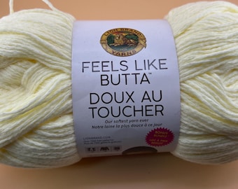 Può includere: Un gomitolo di filato giallo pallido con un'etichetta di carta bianca che riporta il logo Lion Brand Yarns e il testo Feels Like Butta, Doux Au Toucher, Our softest yarn ever, Notre laine la plus douce à ce jour e un cerchio rosa Bonus Bundle.