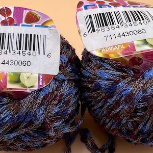 Könnte beinhalten: Zwei Knäuel Garn in einem braunen, blauen und violetten Farbschema. Das Garn ist um einen Pappzylinder mit einem Etikett gewickelt, auf dem "ADRIAFIL" steht und ein Barcode angebracht ist. Die Barcode-Nummer lautet 6 98384 34540 6 7114430060.