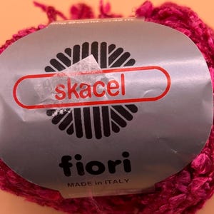 Könnte beinhalten: Knäuel fuchsiafarbenes Garn mit Etikett "skacel fiori MADE IN ITALY". Das Garn ist ein leuchtendes Pink und scheint strukturiert zu sein. Das Etikett ist silberfarben mit schwarzen und roten Akzenten. Für Strick- oder Häkelprojekte.