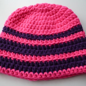 Puede incluir: Un gorro de ganchillo hecho a mano con un vibrante patrón de rayas rosas y moradas. El gorro presenta una corona redondeada y un ajuste ceñido, perfecto para mantener el calor. El gorro tiene una pequeña etiqueta que dice "made by LJ".