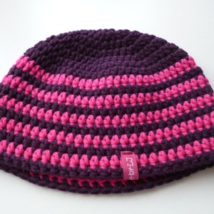 Puede incluir: Un gorro de ganchillo hecho a mano con un patrón de rayas en morado oscuro y rosa brillante. El gorro presenta una textura de tejido apretado y una pequeña etiqueta rectangular con texto. La combinación de colores es vibrante y llamativa.