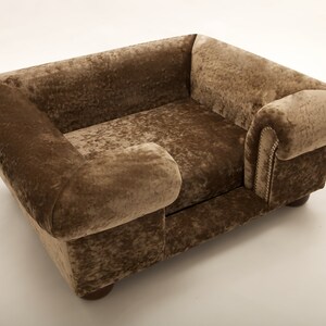 May include: A brown velvet dog bed with a raised headrest and a side armrest. The bed has a tufted design and wooden legs.