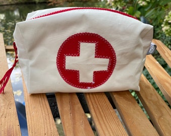 Könnte beinhalten: Weiße Canvas-Kosmetiktasche mit einem roten gestickten Kreuz auf der Vorderseite. Die Tasche liegt auf einer Holzoberfläche.