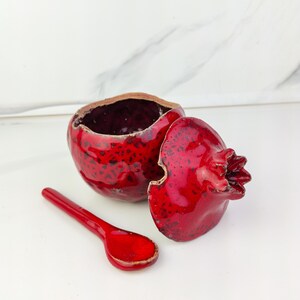 Può includere: Un contenitore in ceramica a forma di melograno con coperchio e cucchiaio abbinati. Il contenitore e il cucchiaio sono di un rosso lucido con macchie scure. Il coperchio è tolto, rivelando l'interno del contenitore.