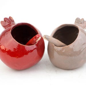 May include: Two ceramic pomegranate-shaped bowls with glossy finishes. One bowl is vibrant red, and the other is a muted taupe. Each bowl features a small, matching ceramic spoon resting inside the open top, which is shaped like a pomegranate crown.