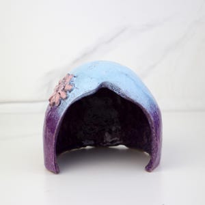 May include: A ceramic hood-shaped sculpture, hand-painted with a gradient of colours. The top is light blue, fading to deep purple at the base. A cluster of pink flowers decorates the side of the sculpture.