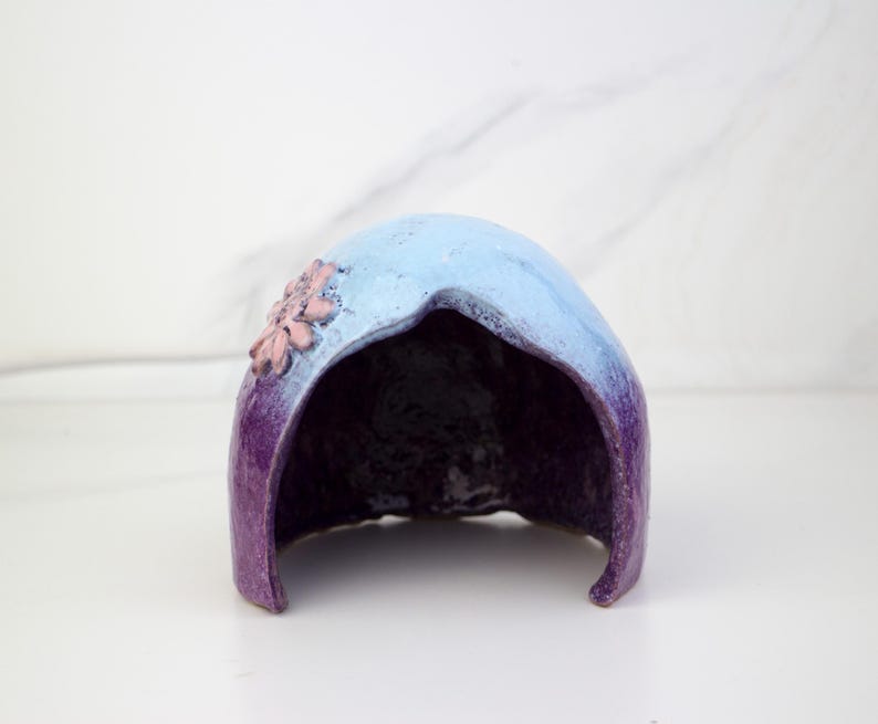 May include: A ceramic hood-shaped sculpture, hand-painted with a gradient of colours. The top is light blue, fading to deep purple at the base. A cluster of pink flowers decorates the side of the sculpture.