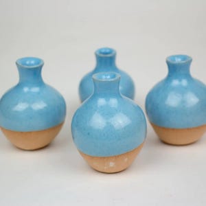 Puede incluir: Cuatro pequeños jarrones de cerámica hechos a mano. Cada jarrón tiene un cuerpo redondeado y un cuello estrecho. La mitad superior es de esmalte azul claro, mientras que la mitad inferior es de color beige natural. Los jarrones están sobre fondo blanco.