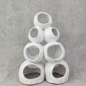 Puede incluir: Una escultura de cerámica blanca compuesta por formas circulares abiertas apiladas. La obra de arte hecha a mano tiene una superficie texturizada y un diseño minimalista. La escultura se exhibe sobre un fondo gris neutro, resaltando sus líneas limpias y su estética moderna.