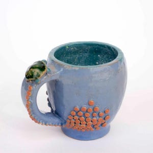 May include: A handcrafted ceramic mug, glazed in blue, featuring an octopus tentacle handle with orange detailing. The mug's interior and rim are teal. A unique, artistic piece for beverages.