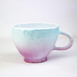 May include: A ceramic mug with a gradient color scheme, transitioning from a light teal at the rim to a soft purple at the base. The mug has a rounded body, a sturdy handle, and a textured surface.