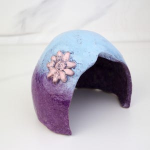 May include: A decorative, cave-shaped object with a light blue top that fades into a deep purple base. A pink flower adorns the front. The interior is a dark purple. This item could be used as a decorative piece or a small animal hideaway.