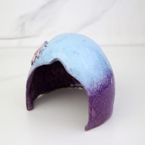 May include: A decorative ceramic piece with a curved form, showcasing a colour gradient. The top is light blue, fading into purple at the base. A small, pink floral detail is visible on the top. The inside is a dark purple colour.