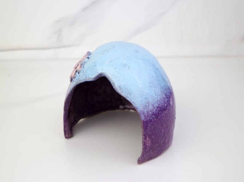 May include: A decorative ceramic piece with a curved form, showcasing a colour gradient. The top is light blue, fading into purple at the base. A small, pink floral detail is visible on the top. The inside is a dark purple colour.