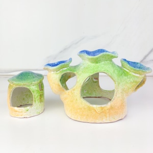 May include: Two ceramic aquarium decorations. One is a small house-shaped structure with a green roof and yellow walls. The other is a larger, coral-like structure with multiple openings, in shades of green, yellow, and blue.