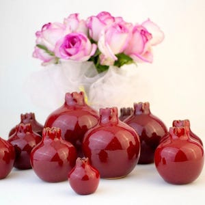 May include: A collection of glossy, deep red pomegranate-shaped vases. The vases are arranged in a group, with a bouquet of pink and white roses in the background. The vases vary in size and have a unique, textured top.