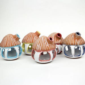 May include: A collection of six ceramic house-shaped candle holders. Each has a brown roof with white vertical lines, a white base, and colorful accents around the openings and chimney. The chimneys are blue, red, and yellow.