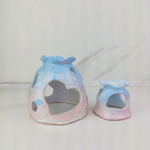 May include: Two ceramic candle holders in a pastel color scheme of pink and blue. The larger holder has a heart-shaped cutout and other circular openings. The smaller holder has a similar design. Both have a ruffled top edge.