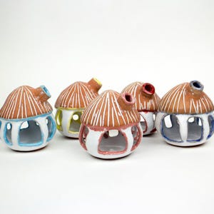 May include: Six ceramic house-shaped candle holders. Each has a brown roof with white vertical lines, a white base, and colorful accents around the windows and chimney. The chimneys are blue, red, yellow, and brown.