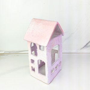 May include: A small, pastel-coloured ceramic house with a pink roof and lavender walls. The house has multiple cut-out windows and arched doorways. The colour gradient transitions from light pink to lavender. The house is a decorative item.
