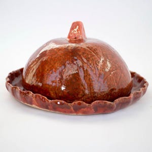 May include: A handmade, ceramic butter dish with a domed lid and scalloped edge base. The dish is glazed in a rich, glossy reddish-brown color. The lid has a small, pointed handle. The dish is designed for kitchen use.