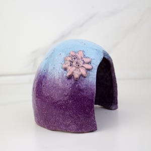 May include: A ceramic, arched structure with a gradient of blue to purple hues. A pink flower adorns the top. The object has an opening in the back, suggesting a decorative or functional purpose.