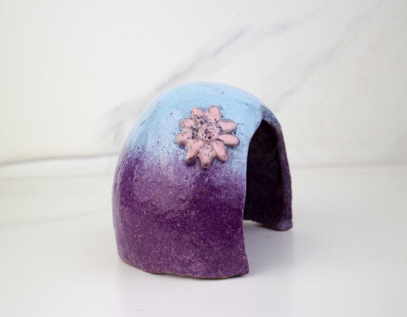 May include: A ceramic, arched structure with a gradient of blue to purple hues. A pink flower adorns the top. The object has an opening in the back, suggesting a decorative or functional purpose.