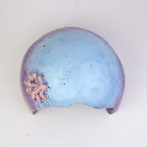 May include: A blue and purple decorative headpiece with a pink flower accent. The headpiece has a textured surface and a curved shape, suitable for a costume or artistic display.