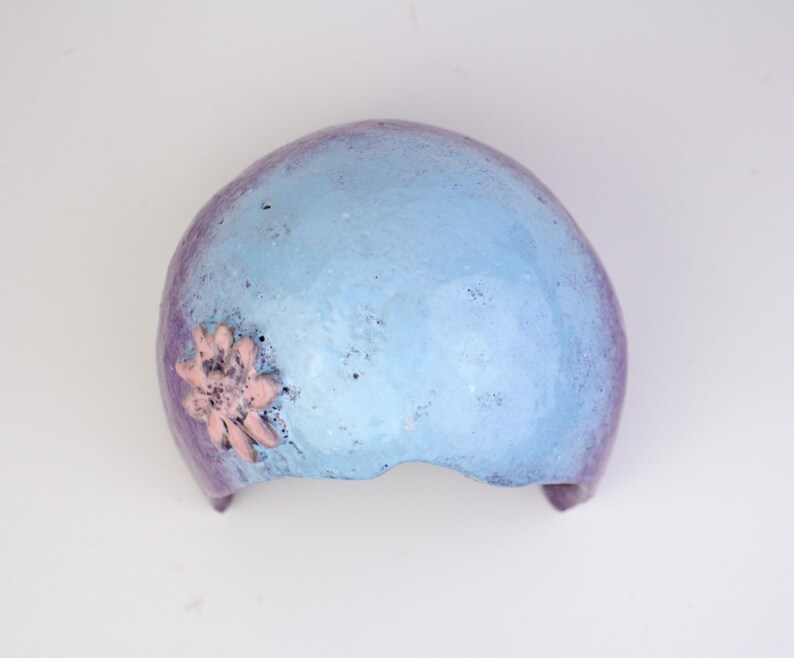 May include: A blue and purple decorative headpiece with a pink flower accent. The headpiece has a textured surface and a curved shape, suitable for a costume or artistic display.