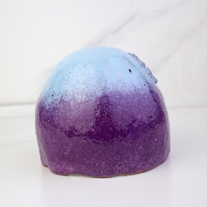 May include: A ceramic sculpture with a rounded form, showcasing a colour gradient. The top is light blue, fading into a deep purple at the base. The surface has a speckled texture, set against a white backdrop. The sculpture is likely handmade.