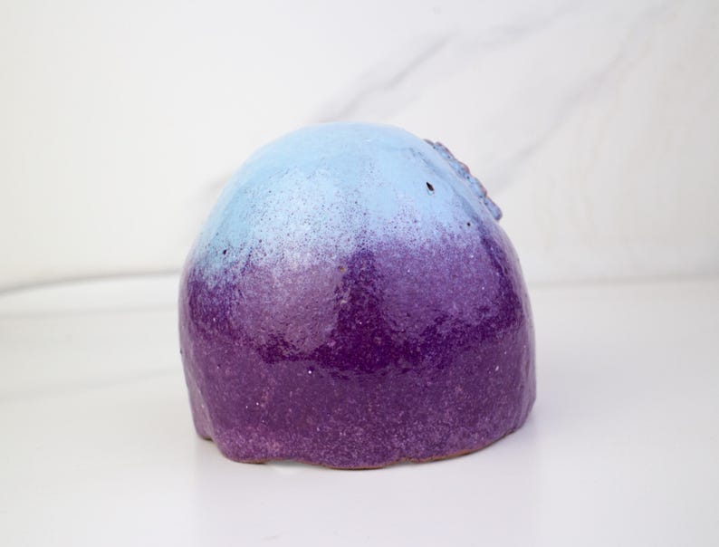 May include: A ceramic sculpture with a rounded form, showcasing a colour gradient. The top is light blue, fading into a deep purple at the base. The surface has a speckled texture, set against a white backdrop. The sculpture is likely handmade.