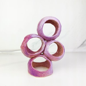 May include: A ceramic sculpture featuring four interconnected circular rings arranged in a vertical cluster. The surface has a glossy, textured finish with a gradient of vibrant purple and pink hues, showing variations in colour intensity throughout.