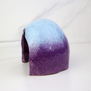 May include: A ceramic sculpture with a rounded arch shape. The top is light blue, transitioning to a deep purple at the base. The object has a textured surface and is set against a white background.