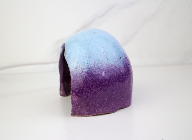 May include: A ceramic sculpture with a rounded arch shape. The top is light blue, transitioning to a deep purple at the base. The object has a textured surface and is set against a white background.