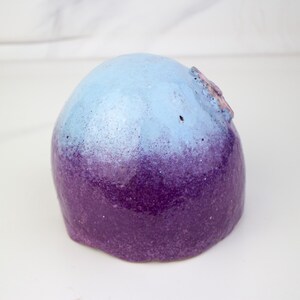 May include: A rounded ceramic sculpture with a colour gradient. The top is light blue, fading into a deep purple at the base. A small, textured detail is visible on the top of the sculpture.