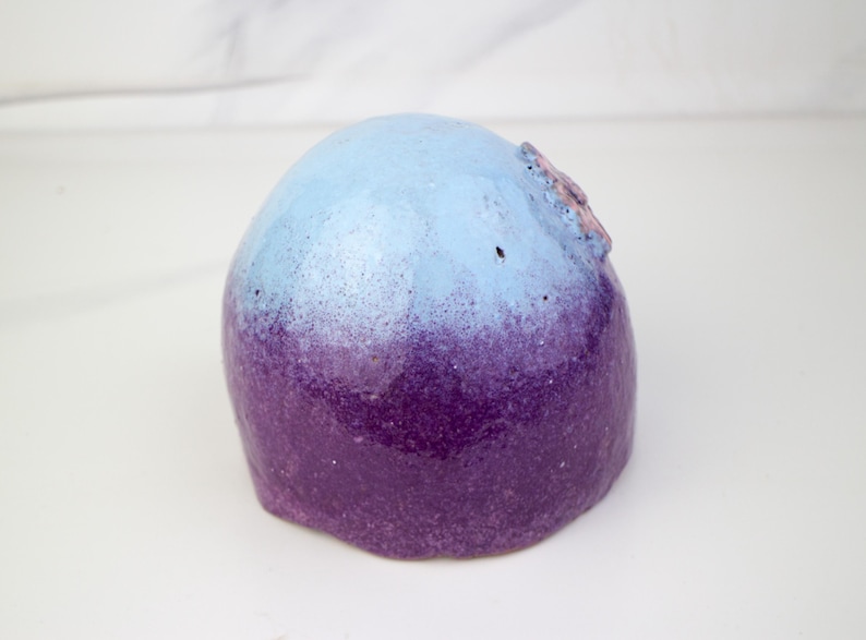 May include: A rounded ceramic sculpture with a colour gradient. The top is light blue, fading into a deep purple at the base. A small, textured detail is visible on the top of the sculpture.