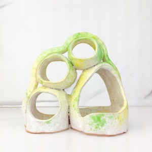 May include: A ceramic sculpture featuring three circular openings and one larger triangular opening. The piece is finished in a white glaze with speckled accents of bright lime green and yellow, creating a textured, organic appearance.