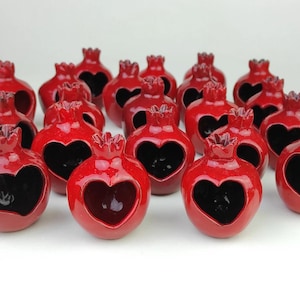 Może przedstawiać: Kolekcja błyszczących, czerwonych ceramicznych rzeźb granatów. Każdy granat ma wycięcie w kształcie serca, odsłaniające ciemne wnętrze. Rzeźby mają teksturowaną koronę.