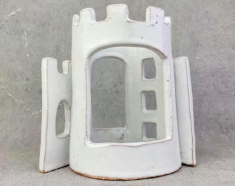 May include: A white ceramic castle-shaped structure featuring a crenellated top, a large arched central opening, and smaller rectangular cutouts on the side walls. The piece has a glossy finish with visible clay edges along the base and openings.