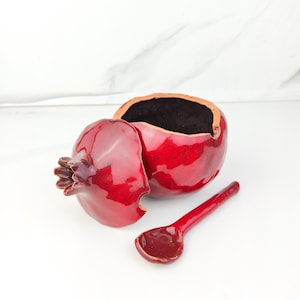 May include: A glossy red ceramic pomegranate-shaped container with a removable lid and a matching small red spoon. The interior of the container is dark, and the exterior features a vibrant, shiny red glaze with a textured, organic fruit shape.