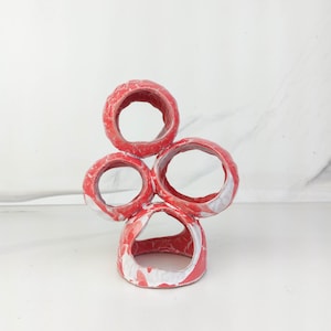 May include: A sculptural object composed of four interconnected circular rings arranged in a vertical cluster. The structure features a marbled finish with vibrant red and white colors, creating a textured, uneven surface across all circular forms.