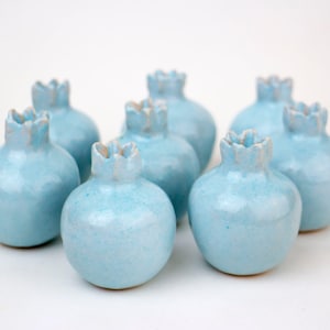 May include: A collection of seven small, light blue ceramic pomegranate-shaped vases. Each vase has a textured, crown-like top and a glossy finish. The vases are arranged on a white surface.
