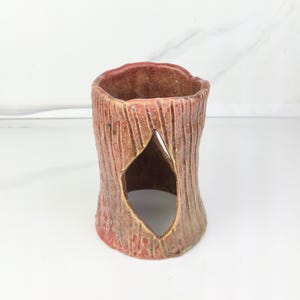 May include: A rustic, tree-trunk-shaped ceramic candle holder. The holder is primarily reddish-brown with textured vertical lines and a diamond-shaped opening. The interior is a darker shade of brown.