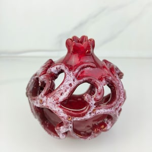 Puede incluir: Objeto decorativo de cerámica roja con forma de granada con intrincados diseños calados. La superficie tiene un acabado brillante con detalles blancos y dorados. La parte superior tiene forma de corona.