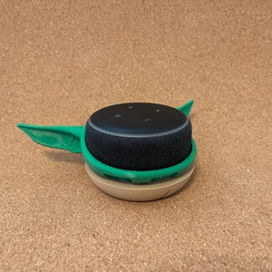 May include: A green and beige 3D printed stand for a black Amazon Echo Dot smart speaker. The stand is shaped like a character from the Star Wars franchise, with two ears sticking out on either side.