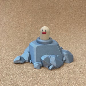 May include: A 3D-printed Diglett figure, a character from the Pokémon franchise. The figure features a beige head with a red mouth and black eyes, emerging from a gray rock formation. The rock has a textured surface.