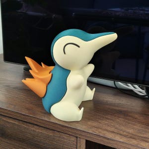 May include: A figurine of a cartoon character with a blue, cream, and orange color scheme. The character has a long beak, a curved black line for a smile, and orange flames on its back. It is sitting on a wooden surface.